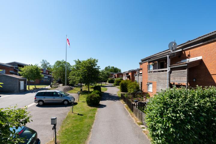 Bolkavägen, två tegelhus, en gångväg, parkering med en bil, grön omgivning och en flaggstång.